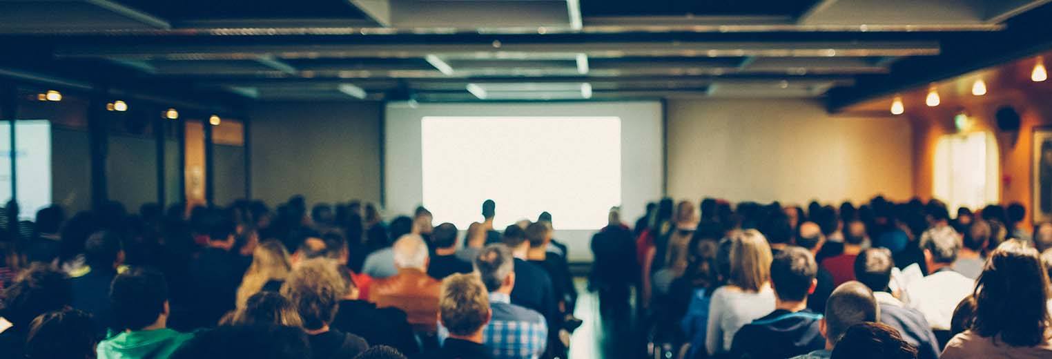 A meeting room with a full room of people sitting down looking forward at a screen. 