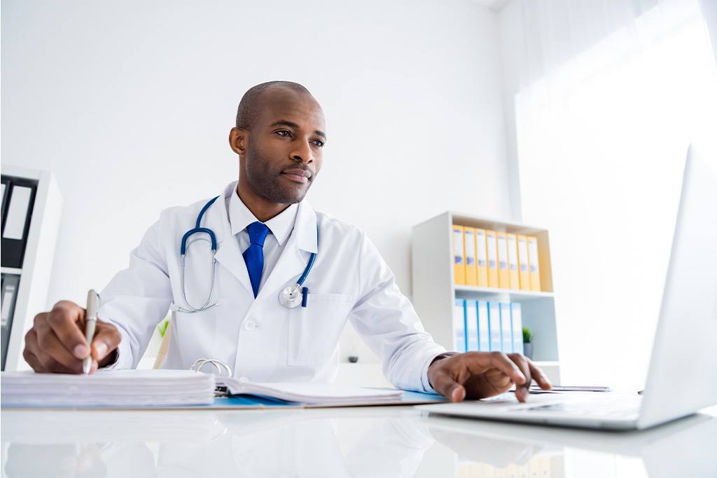 A doctor uses a laptop computer while taking notes on a notepad