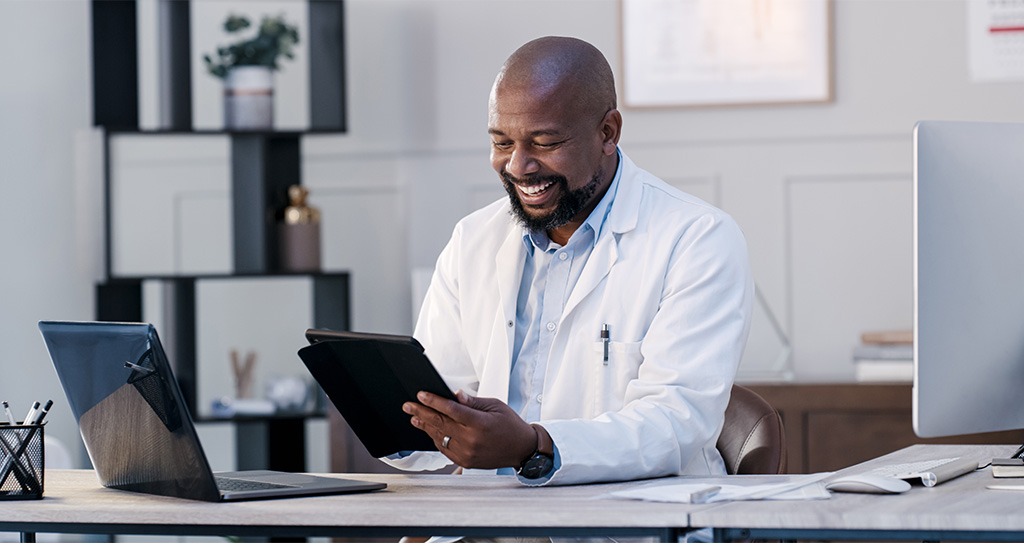A doctor in white lab coat uses a tablet at his desk