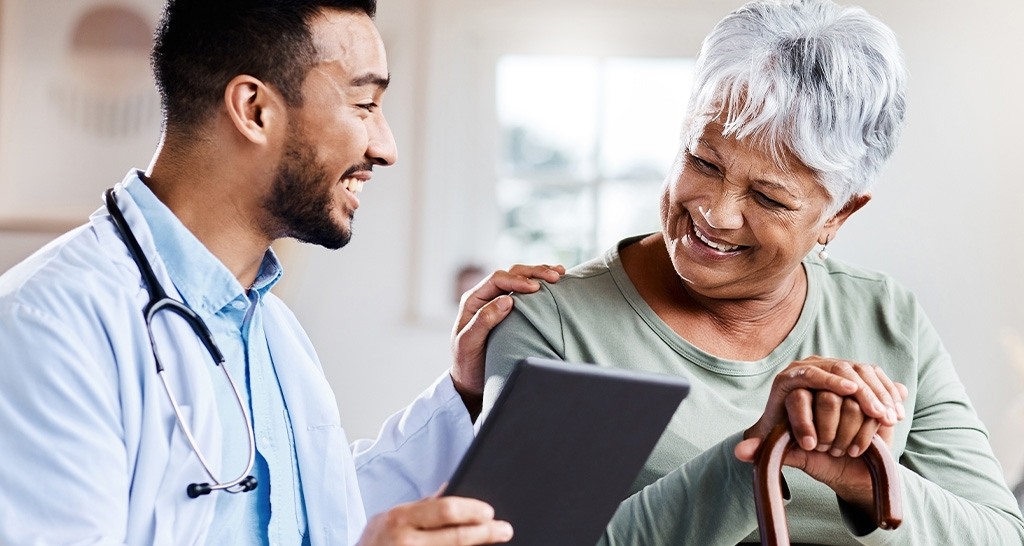 A doctor holding a tablet rests his hand on the shoulder of an elderly patient